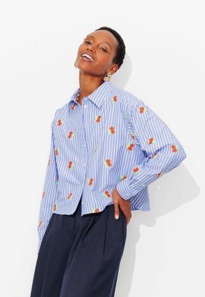 Femme portant une chemise rayée bleu et blanc avec des imprimés floraux orange, un pantalon large à fines rayures foncées, posant avec la main sur la hanche.