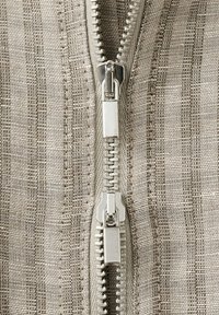 Detailný záber na dvojitý kovový zips na béžovej a hnedej textúrovanej tkanine s vertikálnym pruhovaným vzorom.