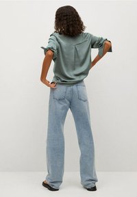 Femme aux cheveux bouclés portant une chemise à manches longues vert sauge et un jean taille haute bleu clair, debout de dos avec les mains sur les hanches.