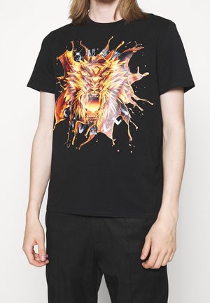 Person, der bærer en sort t-shirt med et flammende geometrisk løvehoved-design og ordene "Just Cavalli" på forsiden.