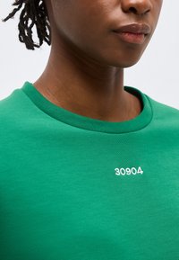 Κοντινό πλάνο ενός ατόμου που φοράει πράσινο πουκάμισο με το "30904" τυπωμένο με λευκά γράμματα στο στήθος, εμφανίζοντας τα χείλη, το λαιμό και μέρος των μαλλιών.