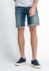 Shorts en denim bleu foncé avec des détails déchirés et des revers. Associés à des chaussures slip-on bleu marine. Matière en coton, design décontracté.
