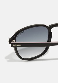 Occhiali da sole in plastica nera con lenti sfumate, sottili accenti metallici e un logo dorato sulla tempia. Presentano un design geometrico moderno.