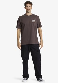Camiseta de algodón marrón oscuro con mangas cortas y un bolsillo en el pecho, que presenta diseños gráficos coloridos. Combinada con pantalones cargo negros holgados.