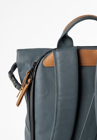 Blaue Rucksack mit glatter Textur, versehen mit einem braunen Lederakzent, robusten Reißverschlüssen und einem oberen Tragegriff.