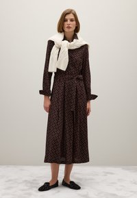 Langes, dunkel gemustertes Kleid mit tailliertem Gürtel, kombiniert mit einem weißen, grob gestrickten Pullover, der über die Schultern gehängt ist, und schwarzen, strukturierten Slippern.