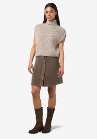 Žena u bezrukavoj pletenoj turtleneck džemperici, smešnom suknji od trakaste tkanine sa dugmadi, i tamnosmešnim čizmama do pola potkolenice koja stoji na beloj pozadini.