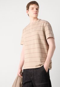 Beige raido t-paita pehmeästä puuvillakankaasta, jossa on lyhyet hihat ja pyöreä kaula-aukko. Hienovarainen logoetuosa rinnassa, yhdistettynä tummiin farkkuihin.