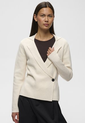 Femme aux longs cheveux foncés porte une veste cache-cœur crème sur un haut sombre et un pantalon noir ample, debout devant un fond clair uni.