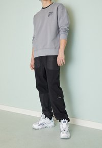 Šedá mikina s černým límcem, která má logo. Černé kalhoty se zipovými kapsami. Bílé tenisky s modrými akcenty a texturovanou podrážkou.