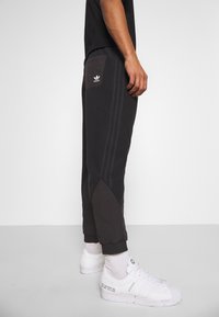 Mörkgrå joggers med fleece-liknande textur, tapered design, sidoränder och en kontrasterande ficka med Adidas-logotypen. Vita sneakers på fötterna.
