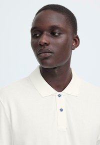 Polo crème en tissu texturé avec col, muni de deux boutons bleus et d'un petit logo sur le côté gauche.