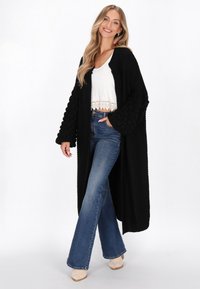 Černý pletený kardigan s texturovanými, oversized rukávy, nošený přes bílý zkrácený top a v kombinaci s rozšířenými džíny.