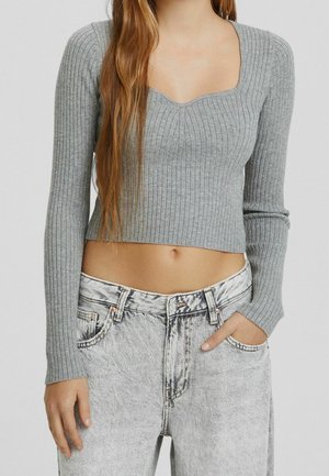 Kobieta ubrana w szary, prążkowany, długi rękaw, krótki top i wysokiej talii jeansy w stylu acid-wash, z jedną ręką w kieszeni i długimi włosami spływającymi na ramię.