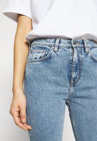 Jean en denim bleu clair avec taille haute, fermeture à boutons, design à cinq poches, coutures contrastantes et finition légèrement délavée.