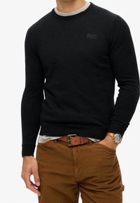 Maglione nero lavorato a maglia con maniche lunghe e scollo tondo, indossato sopra una camicia grigia chiara. Pantaloni in velluto a coste marrone e una cintura marrone.