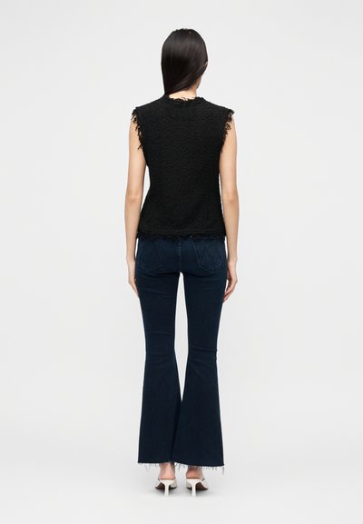 Top preto sem mangas com tecido texturizado e bordas frisadas, combinado com jeans escuros de corte flare e saltos brancos. Modelo mostrado de costas.