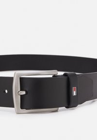 Zwarte leren riem met een zilveren rechthoekige gesp en een klein merklogo-label in rood, wit en blauw. Glad oppervlak, klassiek ontwerp.
