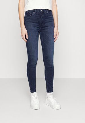 Femme portant un jean skinny bleu foncé, des chaussettes blanches et des baskets blanches, debout devant un fond gris clair.