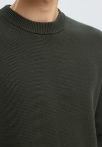 Primo piano di una persona che indossa un maglione lavorato a maglia verde scuro con scollo rotondo, che mostra la texture e i dettagli del colletto.