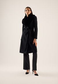 Femme portant un long manteau noir ceinturé avec un grand col en fourrure, un pantalon noir et des chaussures noires à talons hauts, debout devant un fond uni.