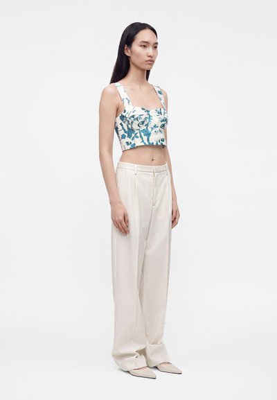 Γυναίκα φοράει ένα λευκό cropped top με μπλε φλοράλ σχέδιο, σε συνδυασμό με άνετο, ψηλόμεσο κρεμ παντελόνι και μυτερές μπεζ γόβες, στέκεται πλαγίως.
