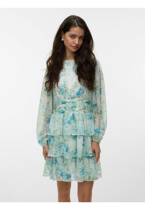 Femme aux longs cheveux foncés portant une robe transparente à manches longues et à volants avec un imprimé floral bleu sur un fond clair.