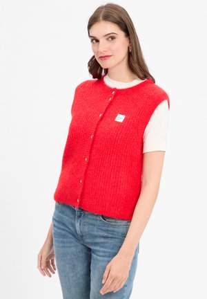 Vrouw in een felrode gebuttonde gebreide vest over een wit T-shirt met korte mouwen en blauwe jeans, tegen een effen witte achtergrond.