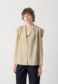 Lys olivengrønn bluse med et mykt, drapert design, korte ermer og knappestolpe foran, kombinert med mørk marineblå bukser.