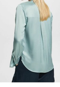 Ljus teal satenblus med rundad nederkant, en bakfåll och knäppte manschetter. Tyget har en slät, glansig yta.