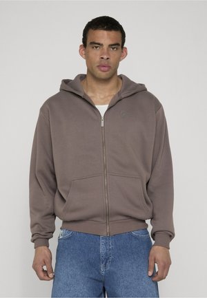 Bărbat purtând un hanorac maro cu fermoar și buzunare frontale, cu o cămașă albă dedesubt, asortat cu blugi albaștri, pe un fundal simplu.