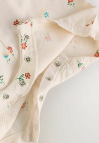 Chemise de couleur crème avec des motifs floraux rouges, bleus et verts. Présente des fermetures à pression et un tissu doux et texturé.