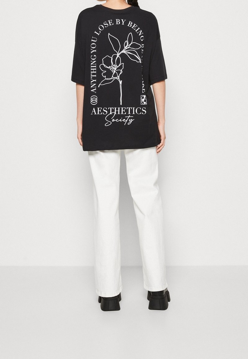 Personne portant un t-shirt noir oversize avec un motif floral blanc et un texte dans le dos, un pantalon blanc et des chaussures noires, de dos à la caméra.