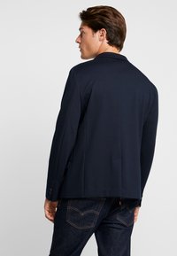 Blazer navy de corte ajustado, fabricado en tejido suave. Presenta dos botones, solapa con muesca y una abertura en la parte posterior. Combinado con vaqueros oscuros.