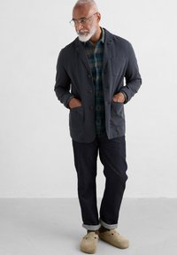 Blazer dintr-un material gri închis, cu guler tip șal, buzunare frontale, purtat peste o cămașă cu carouri verzi, asortat cu pantaloni din denim închis și saboți bej.