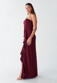 Burgundy, pitkä mekko yhdellä olkaimella, röyhelöhelma ja reiden korkeudelle ulottuva halkio. Sileä rakenne ja istuva yläosa.