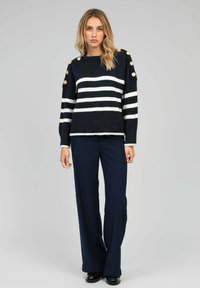 Pull rayé bleu marine et blanc avec un large col rond, tricot texturé et boutons décoratifs sur les épaules. Associé à un pantalon large bleu marine.