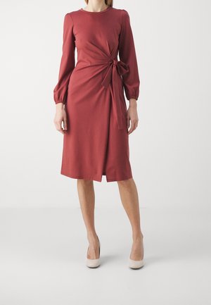 Mujer que lleva un vestido rojo óxido de manga larga y longitud hasta la rodilla con un lazo lateral y tacones altos beige, de pie frente a un fondo blanco.