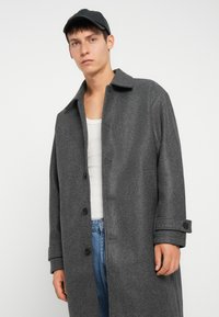 Grå ullfrakk med krage, frontknapper og oppbrettede ermer, kombinert med en hvit tanktop og blå jeans.