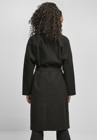 Lang svart frakk med brede ermer, belted midje og en glatt tekstur, med rett snitt og minimalistisk design.