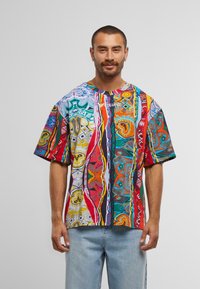 Mann mit kurzen Haaren und Bart, der ein mehrfarbiges, abstrakt gemustertes T-Shirt und hellblaue Jeans trägt, steht vor einem einfarbigen Hintergrund.