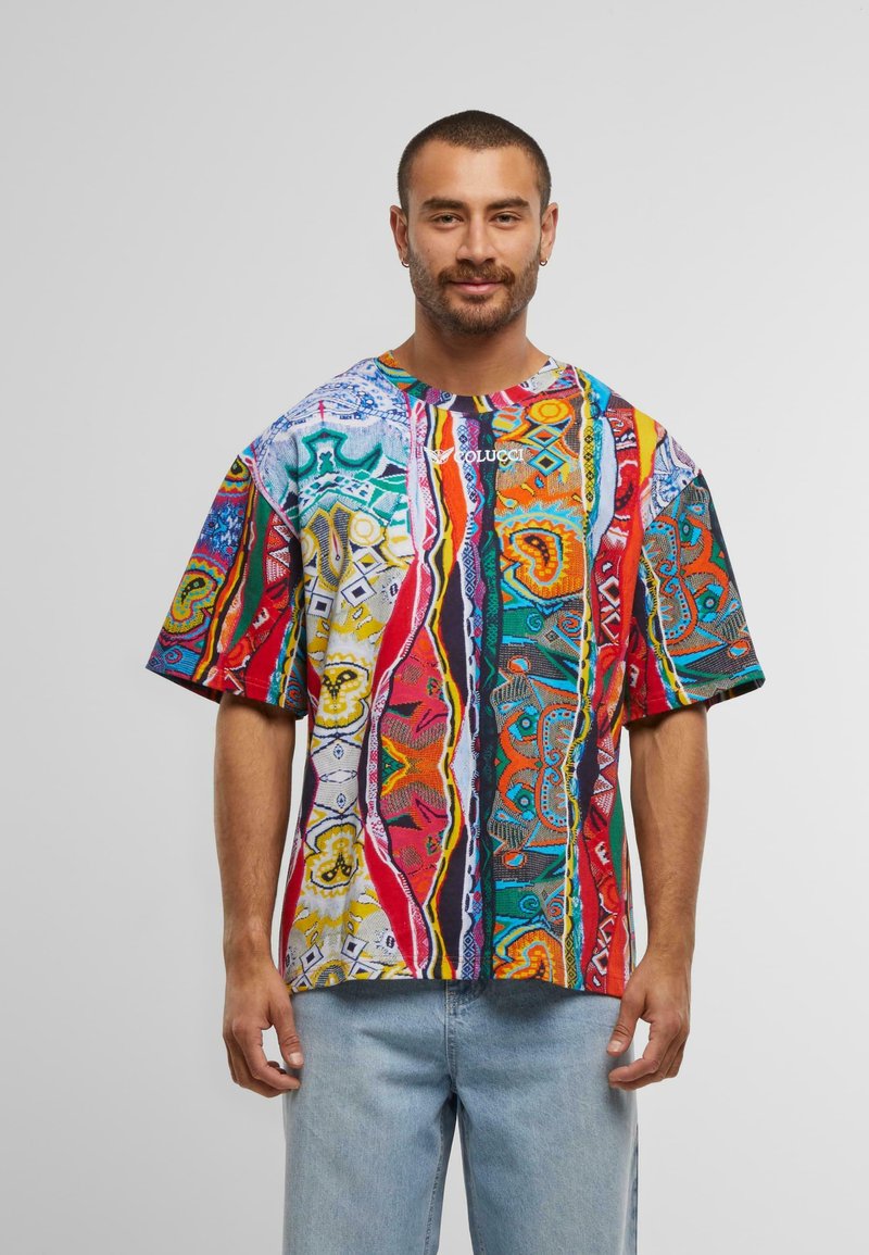Hombre con cabello corto y barba, vistiendo una camiseta de diseño abstracto multicolor y jeans de color azul claro, de pie contra un fondo liso.