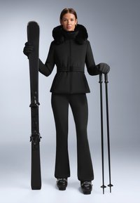 Fekete síruha, prémmel díszített gallérral, testhez simuló dzsekivel, övvel, bővülő nadrággal, sílécekkel és botokkal állva.