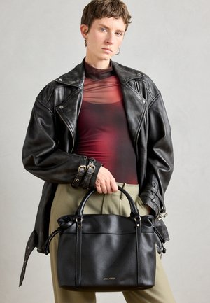 Melnas akmenti ādas somiņas ar divām rokturām, ar zelta furnitūru un vienkāršu, strukturētu dizainu. Modelis valkā melnu ādas jaku.