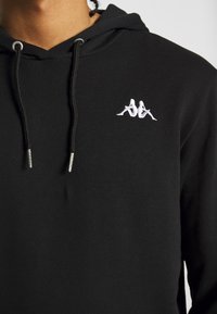 Gros plan d'un sweat à capuche noir avec un cordon et un petit logo blanc représentant deux figures assises dos à dos sur la poitrine.