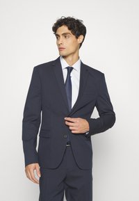 Marineblaues, tailliertes Sakko mit zwei Vorderseiten-Taschen, einem Ein-Knopf-Verschluss und Reverskragen. Getragen über einem hellblauen Hemd und einer dunklen Krawatte.