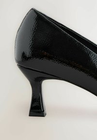 Scarpa con tacco alto in pelle verniciata nera, con punta affusolata e tacco stretto e quadrato, su uno sfondo chiaro.