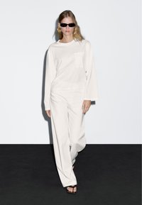 Weißes Langarmoberteil mit Tasche, kombiniert mit weißen weiten Hosen. Das Model trägt schwarze Sonnenbrillen und braune Sandalen vor einem dunklen Hintergrund.