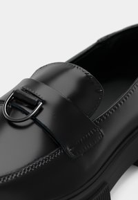 Black leather loafers featuring a rounded toe, sleek finish, decorative strap, and a silver-tone buckle accent. Textured stitching detail visible.