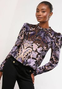 Caurspīdīga melna blūze ar purpura un zelta ziedu rakstiem, savāktā apkakle un pūkainas garās piedurknes; apvienota ar melniem biksēm ar zelta detaļām.